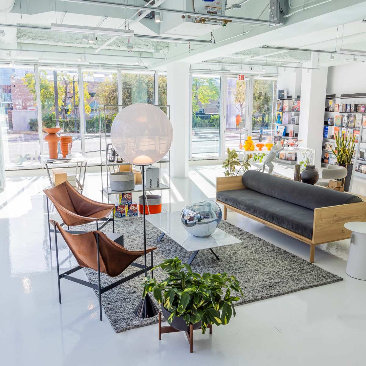 An open, modern living space features a combination of sleek furniture, a large circular lamp, and shelves displaying books and decorative items, all illuminated by natural light.