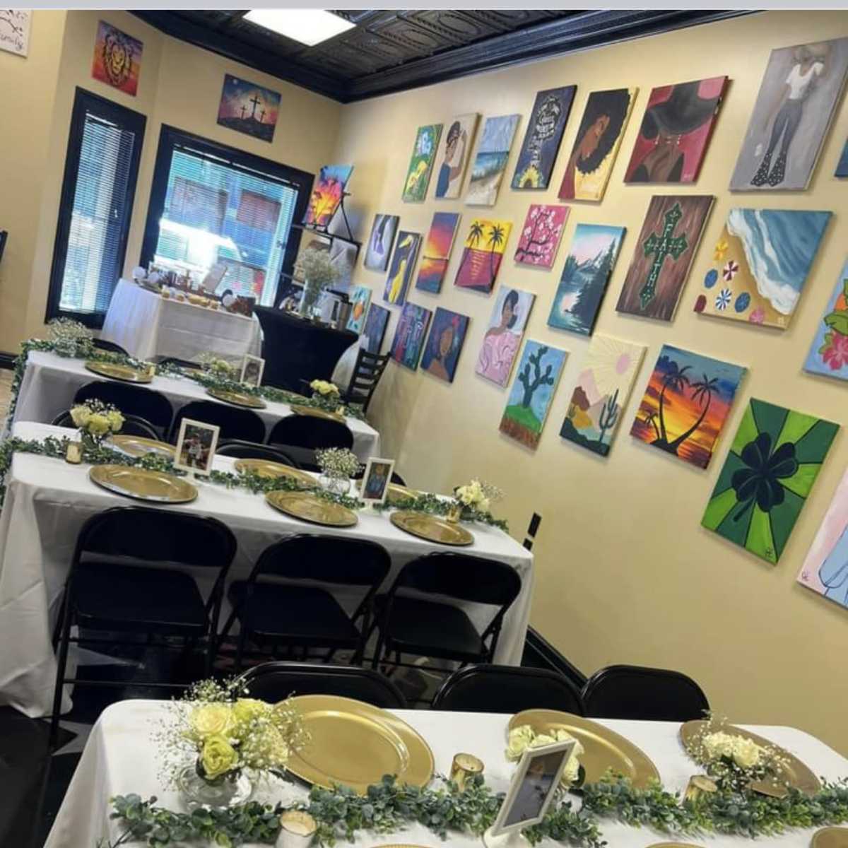 A decorated dining area with multiple tables set for an event, featuring gold plates and floral centerpieces against a backdrop of colorful artwork on the walls.