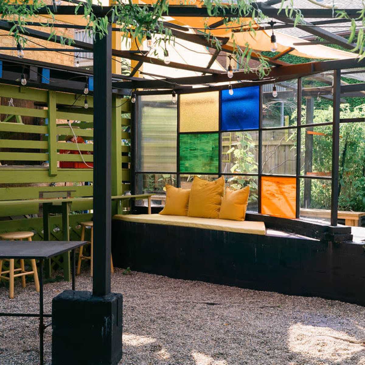 A modern outdoor seating area features a large, colorful glass panel wall, a yellow cushioned bench, and gravel flooring surrounded by greenery.