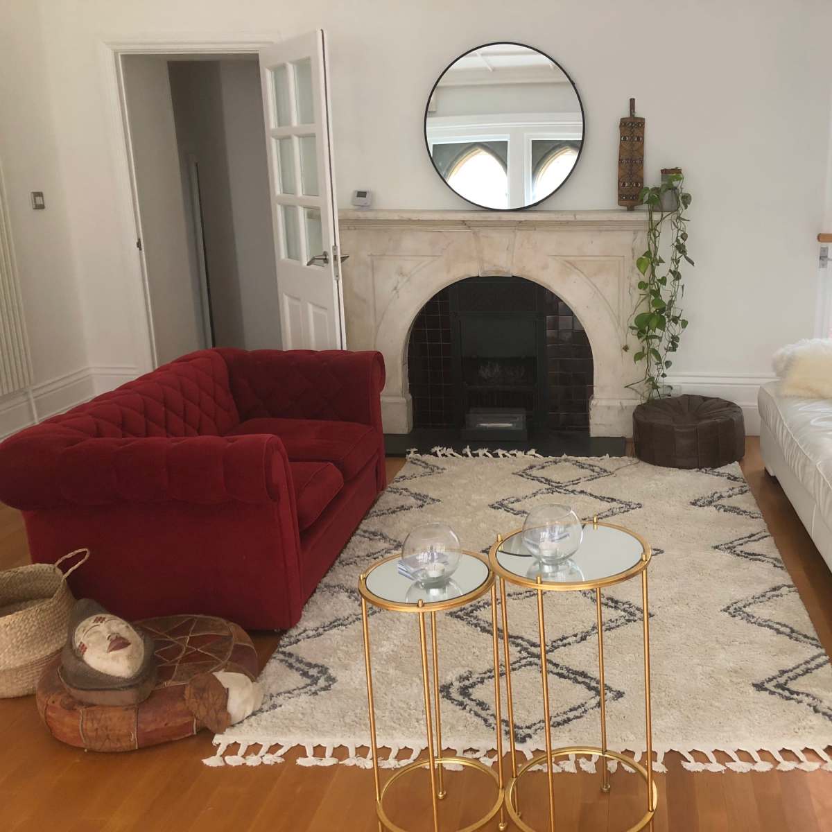 A living room with a red velvet sofa, a white leather couch, a marble fireplace, and two gold side tables on a patterned area rug.