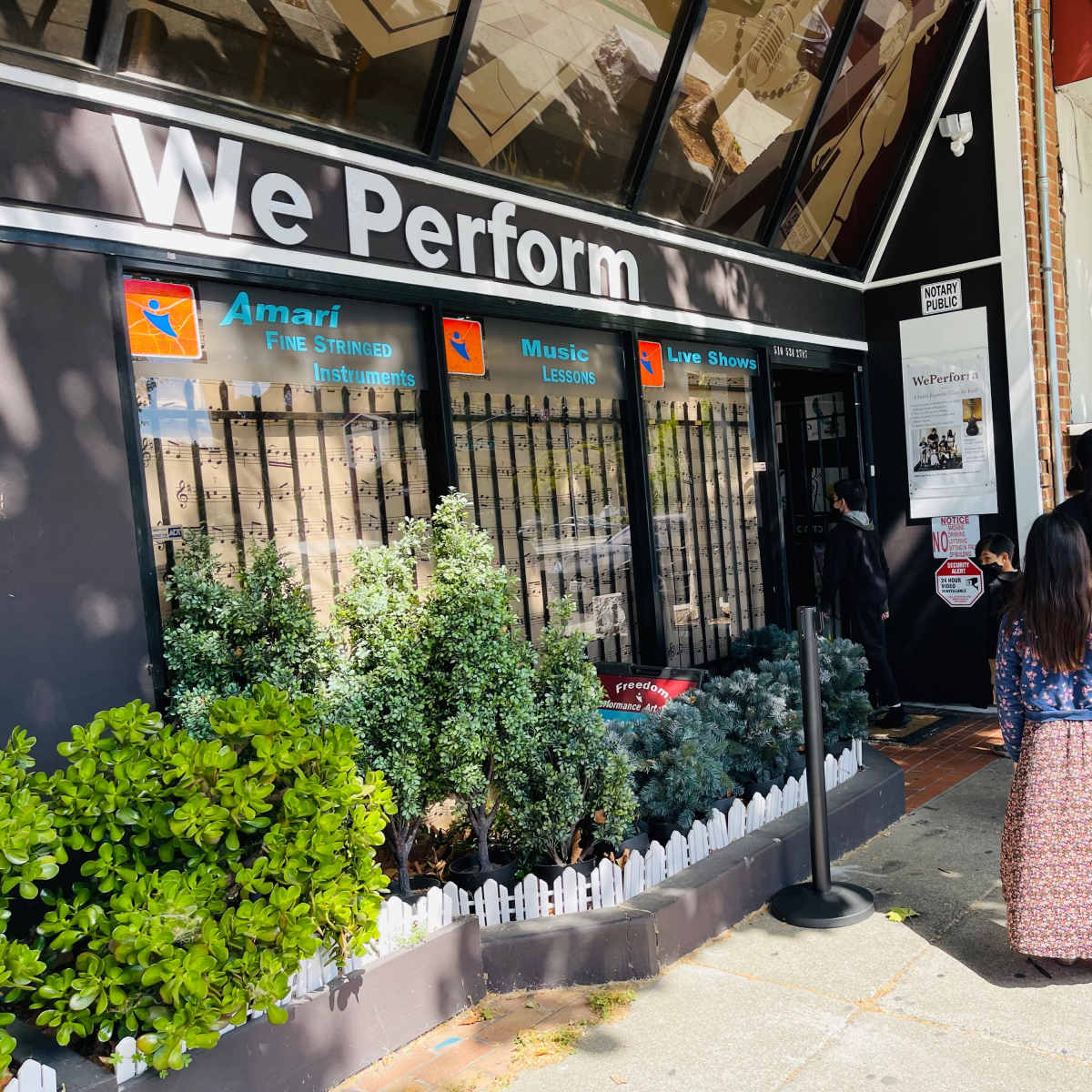 A woman and a child stand in line outside a building labeled "We Perform," which features a sign indicating music and live shows.