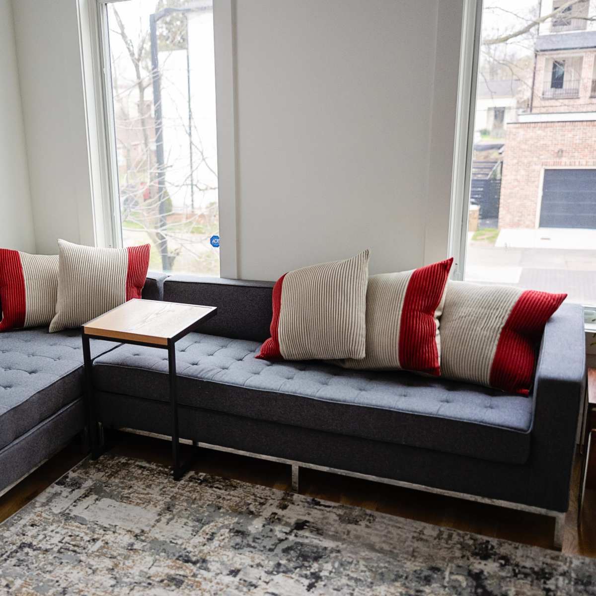 A gray sectional sofa with red and beige pillows is positioned next to a small wooden table in a well-lit room with large windows.