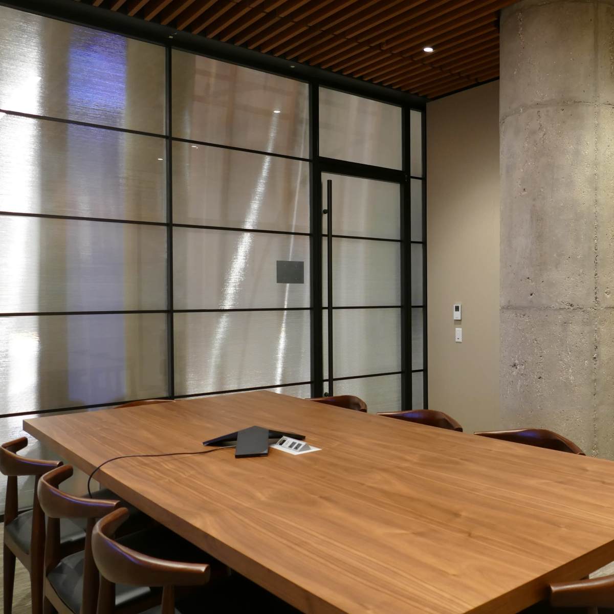 A modern meeting room features a wooden table surrounded by chairs, a concrete pillar, and a glass wall leading to another space.