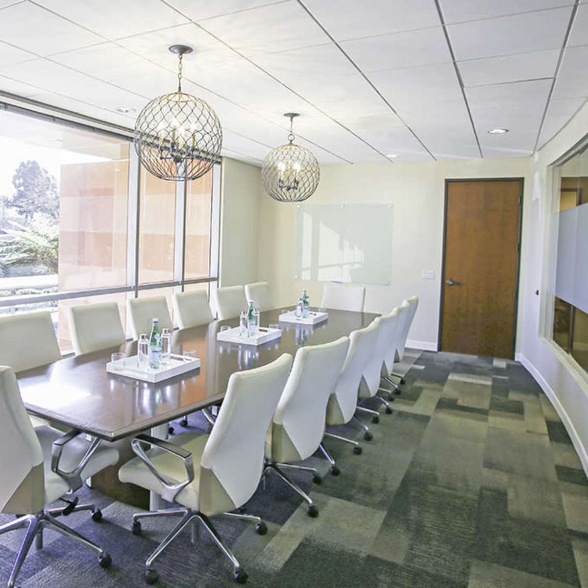 A modern conference room with a long table surrounded by chairs, large windows allowing natural light, and decorative light fixtures hanging above.