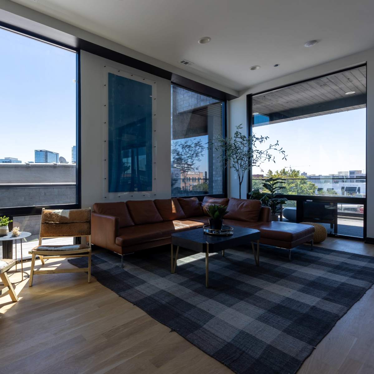 A modern living room features a brown leather sofa, two wooden chairs, a coffee table, and large windows with an urban skyline visible outside.