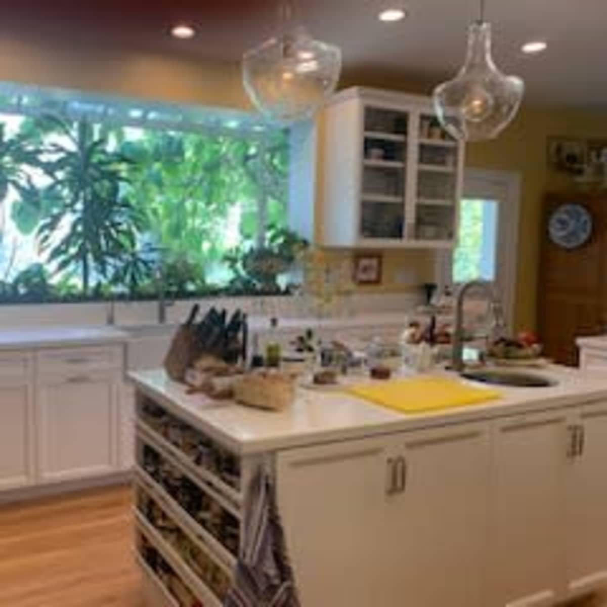 A bright kitchen features white cabinetry, a central island with storage, and large windows displaying lush green plants.