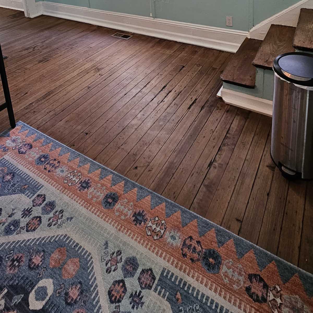 A room with light blue walls, two windows, a wooden floor, and a staircase leading to a lower area.