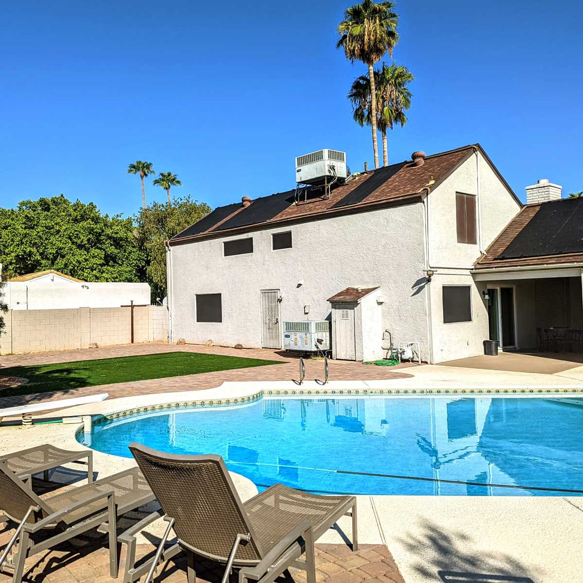 A swimming pool with lounge chairs is situated in the foreground, with a two-story house and palm trees in the background under a clear blue sky.