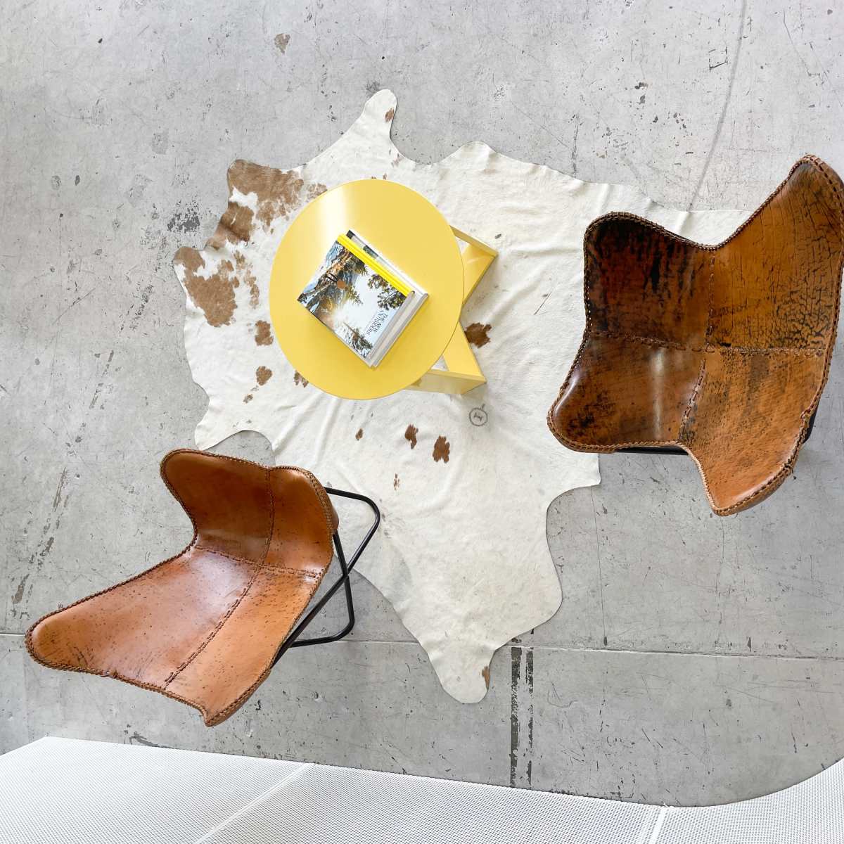 Two brown chairs with a yellow side table are arranged on a white cowhide rug in a minimalist space.