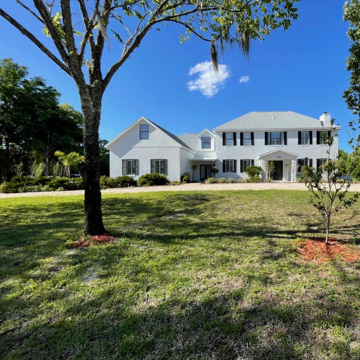 A two-story white house with black shutters is set in a grassy yard surrounded by trees.
