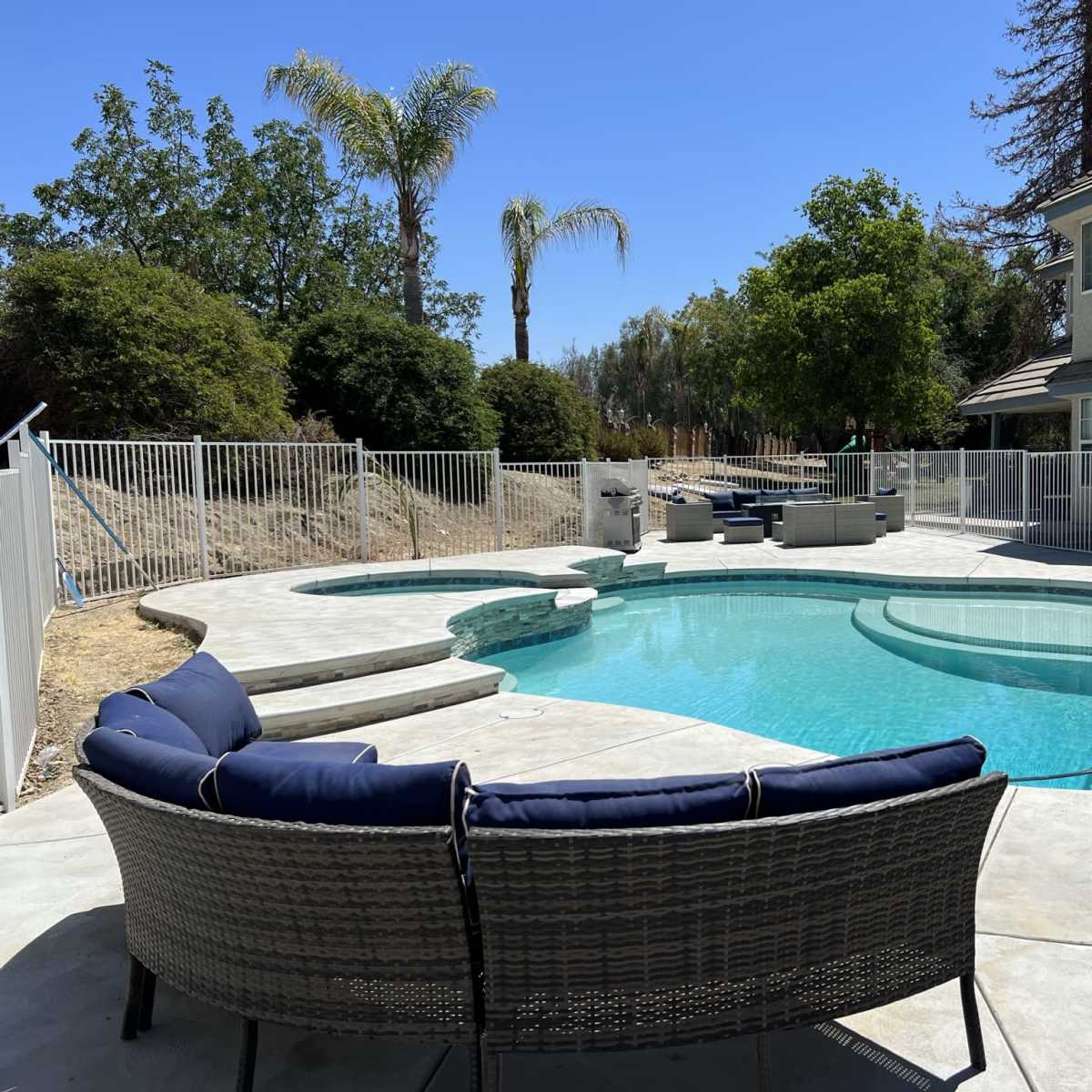 A curved swimming pool surrounded by a concrete patio and a white fence, with outdoor seating and palm trees in the background.