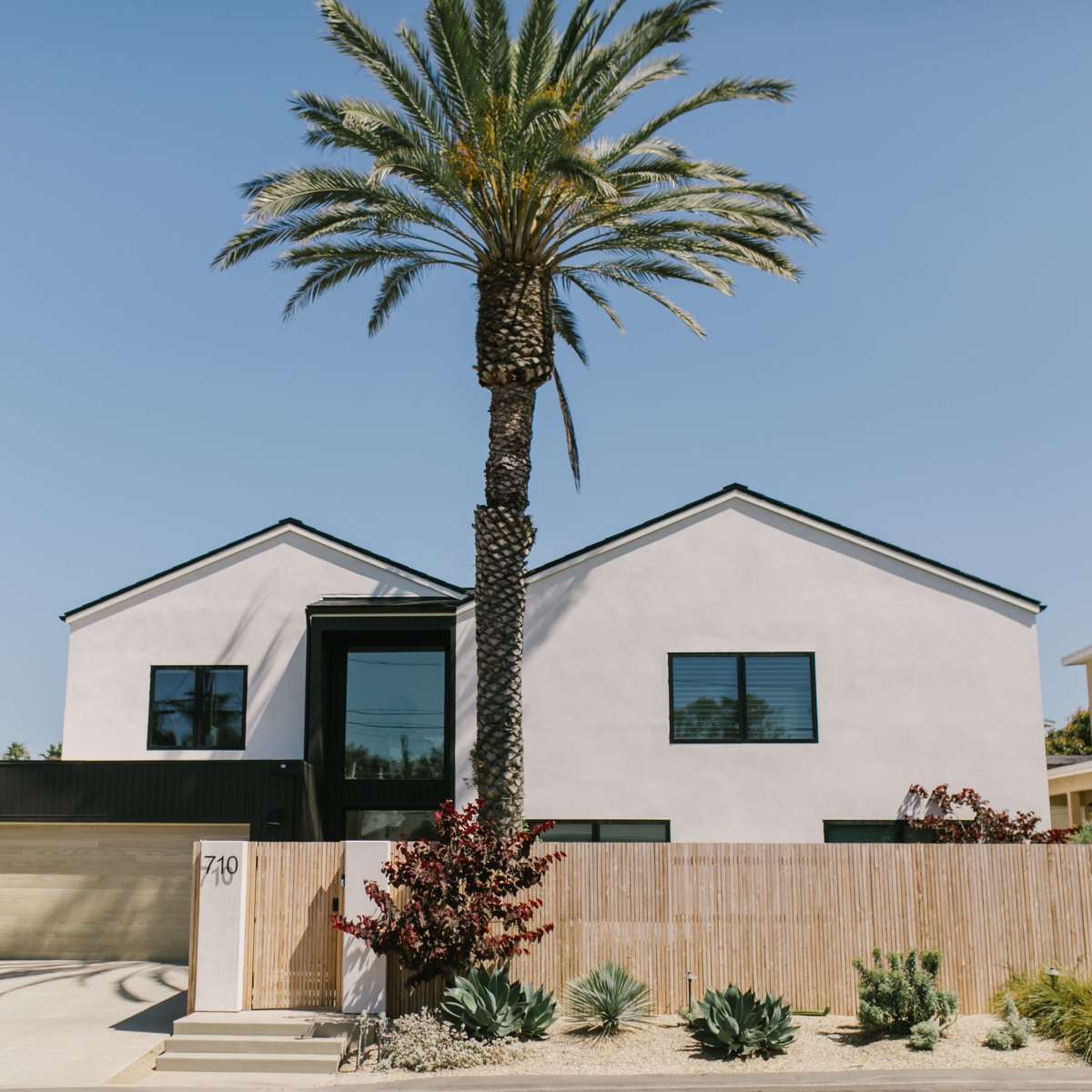A modern two-story house with a black roof and large windows is situated next to a tall palm tree and surrounded by a low fence.