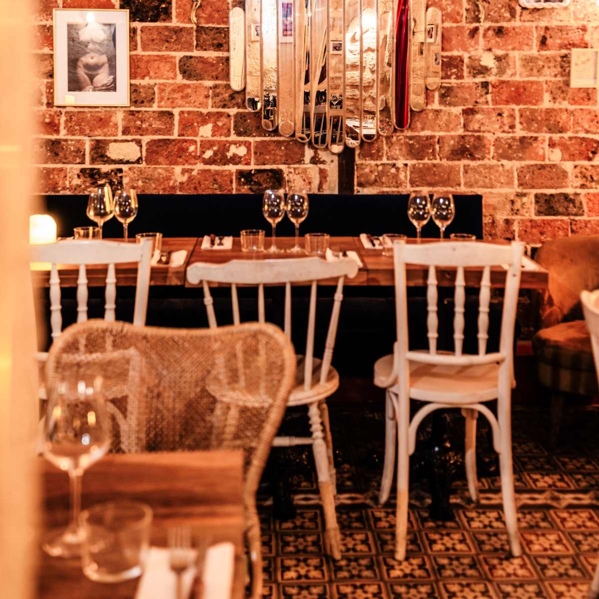A rustic restaurant features a brick wall adorned with a decorative mirror and framed photographs, alongside wooden tables set for dining.