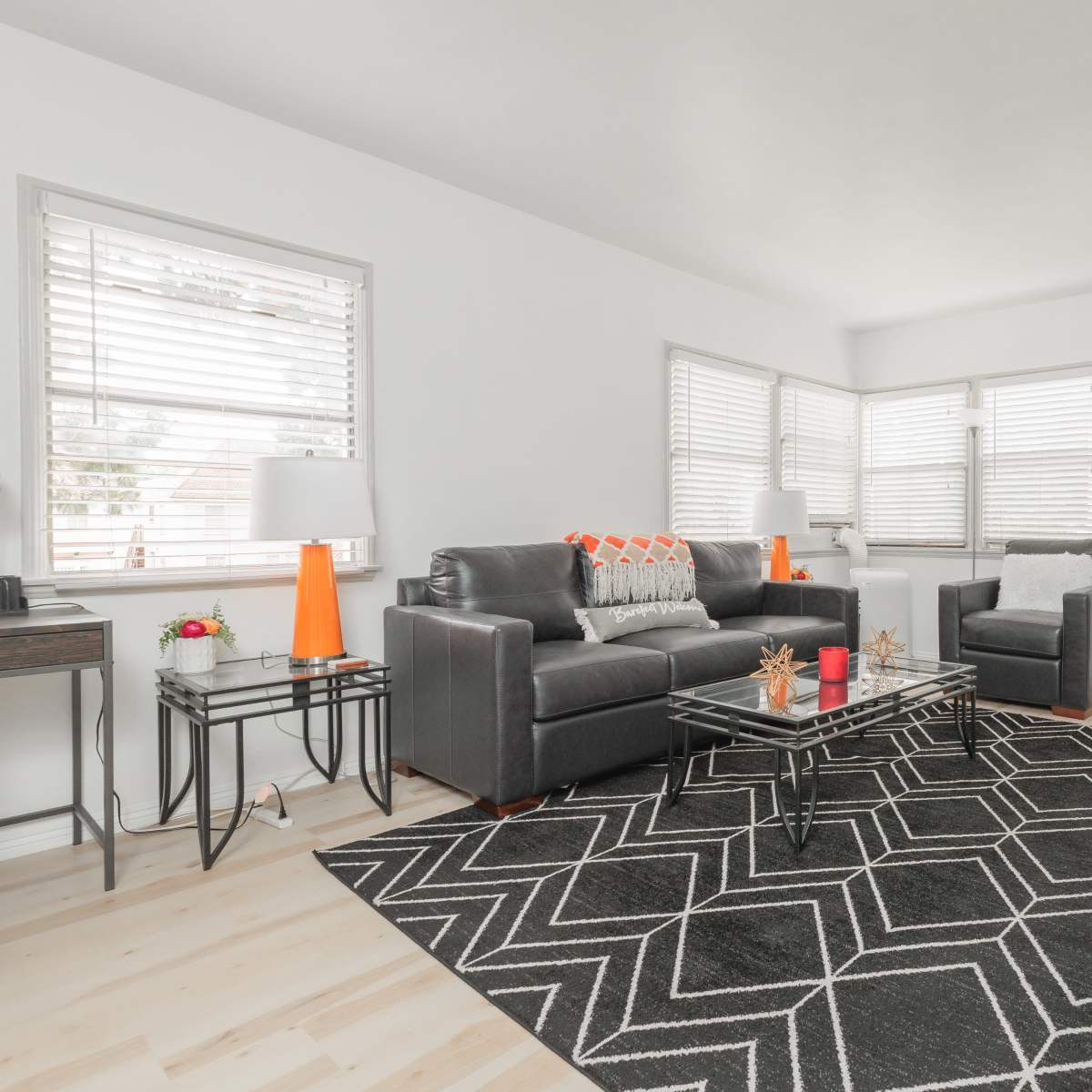 A modern living room features a black and white geometric rug, a glass coffee table, and a gray seating arrangement with bright accents.