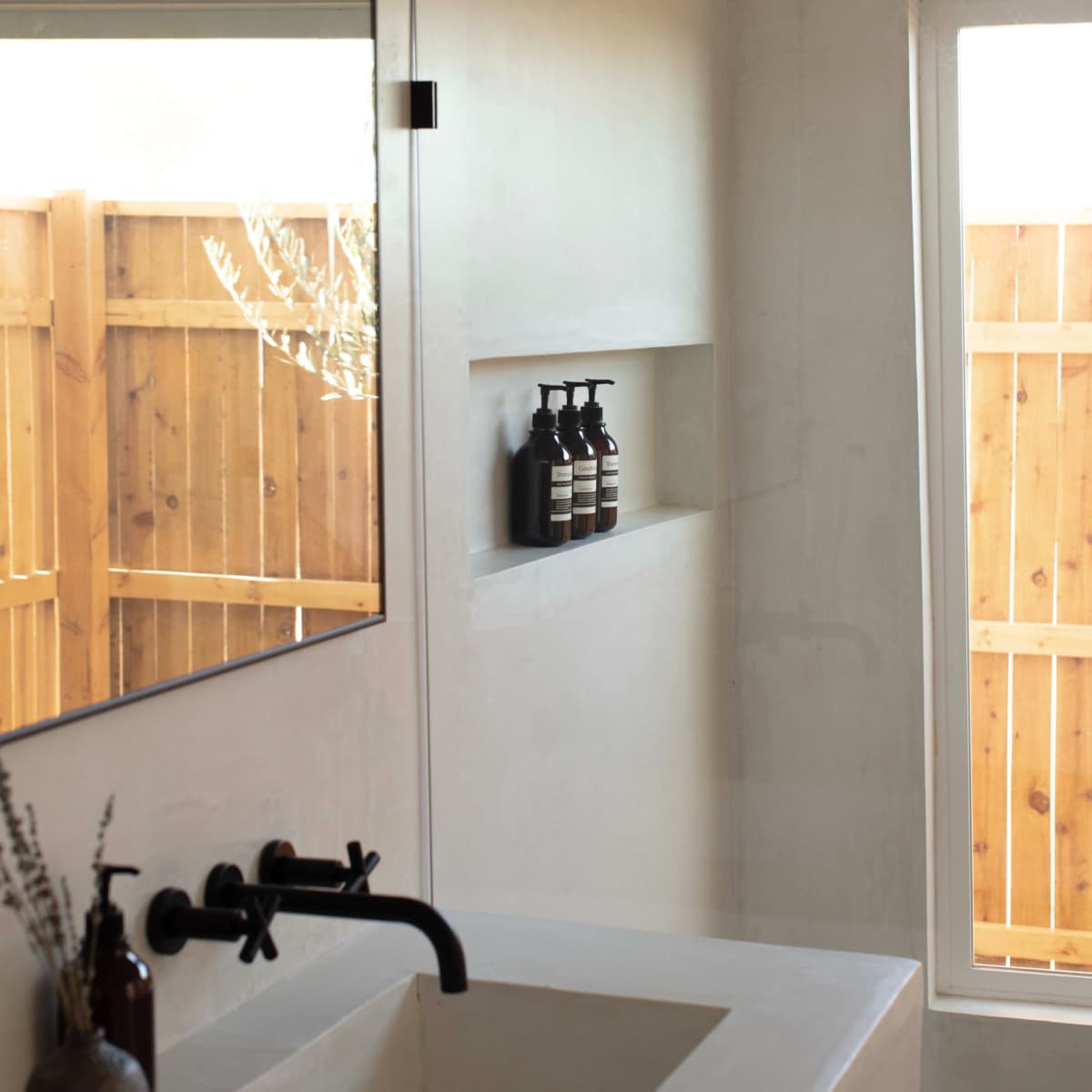 A modern bathroom features a large mirror, a minimalist sink, and a niche displaying dark bottles, with a wooden fence visible through the window.