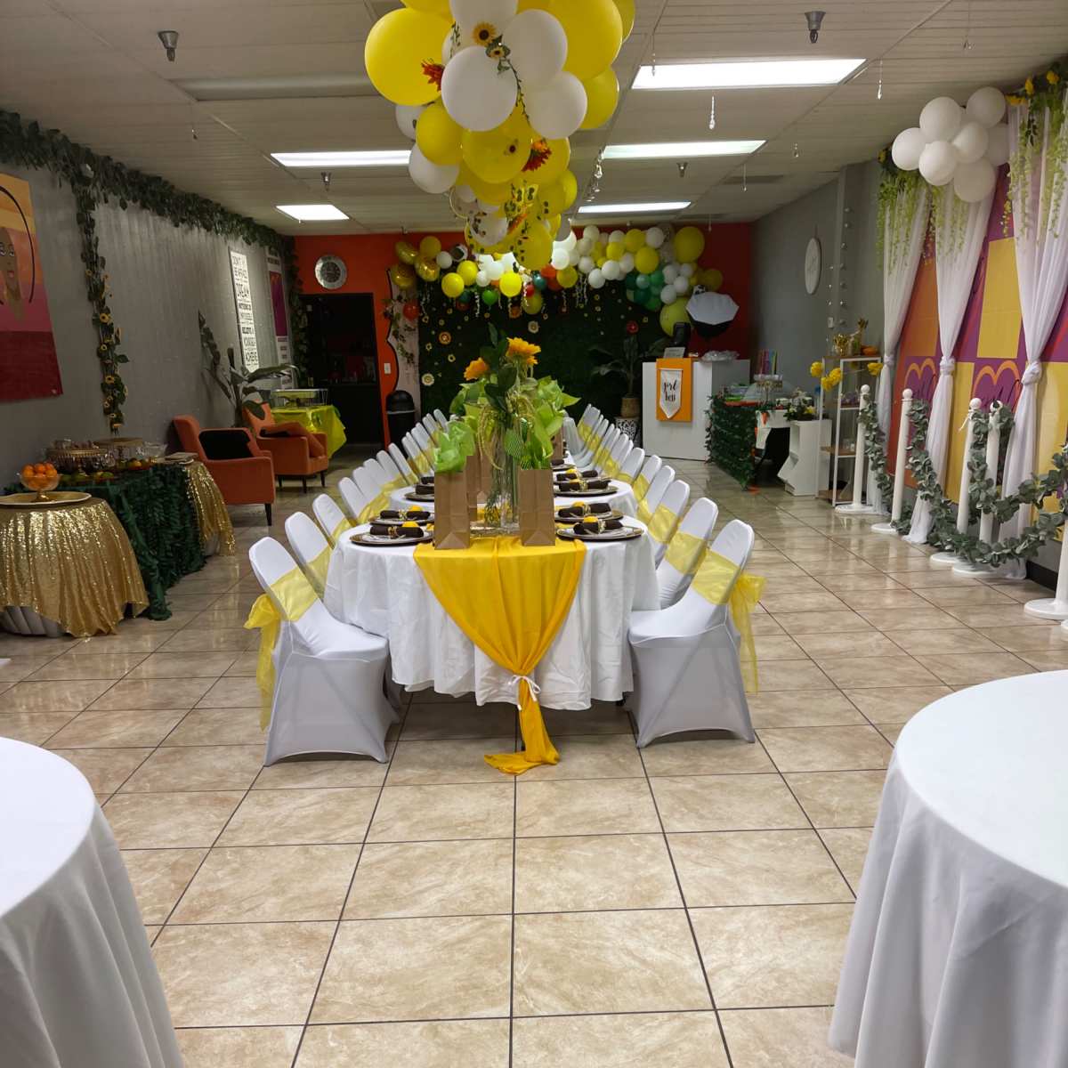 A decorated banquet hall with a long table set for an event, featuring white chairs and yellow accents, surrounded by balloons and floral arrangements.