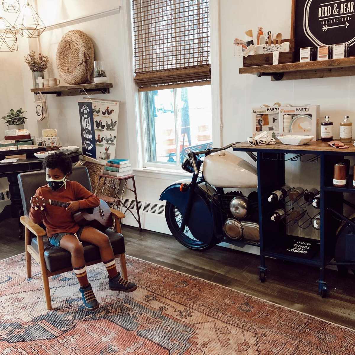 A child sits on a chair playing a ukulele in a cozy shop filled with home decor and a vintage motorcycle.
