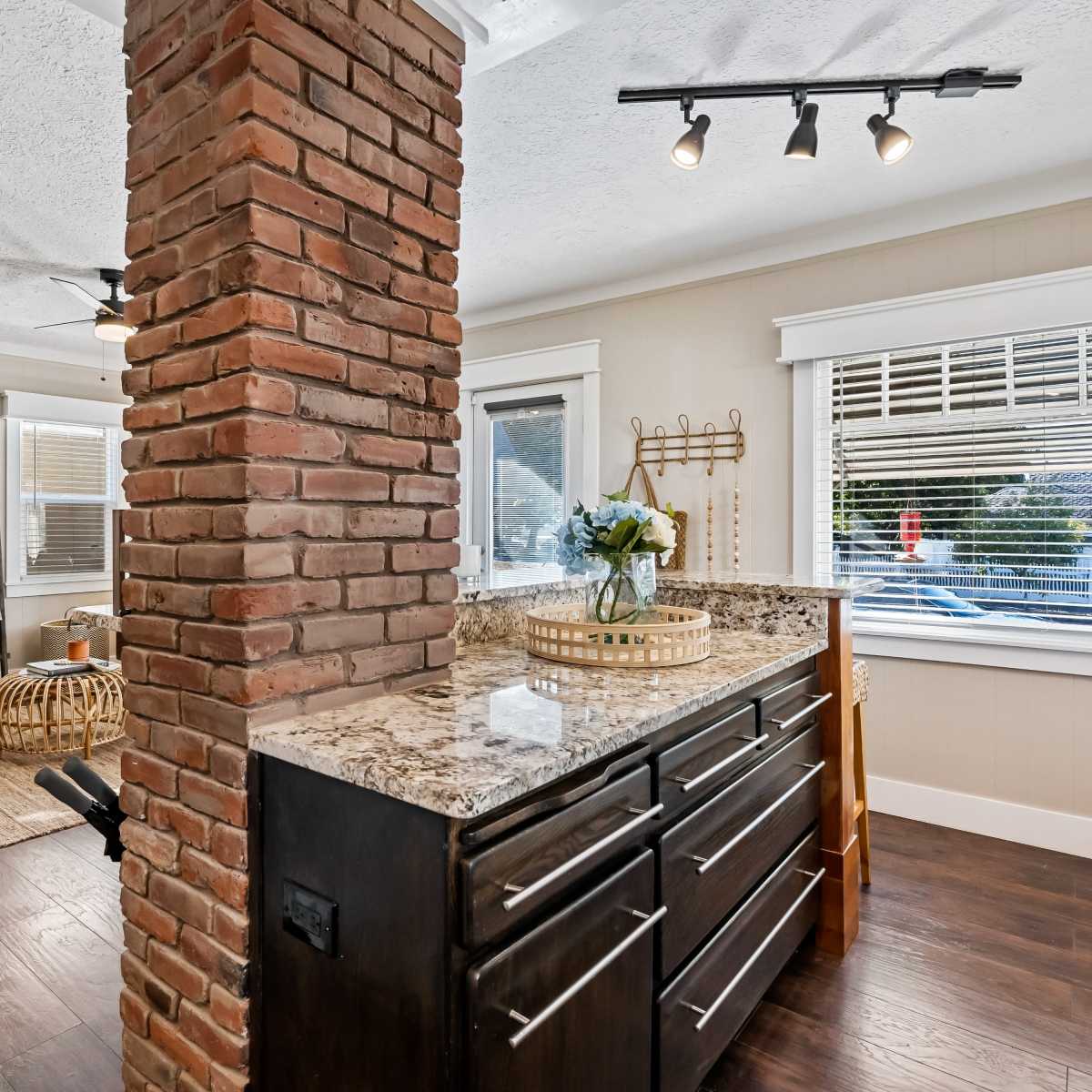 A modern kitchen with a granite countertop and a brick column separating it from a cozy seating area featuring wicker furniture and large windows.
