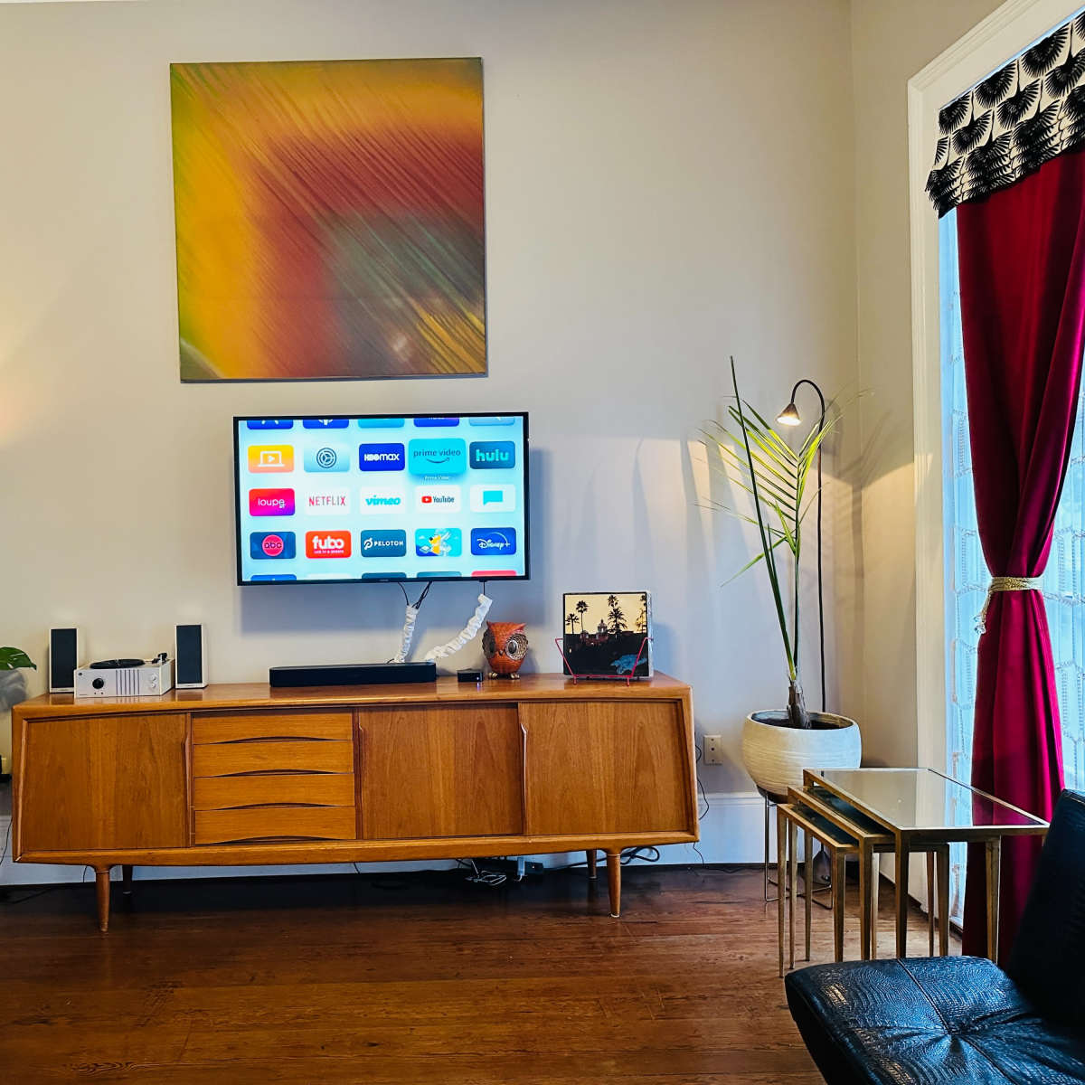 A living room features a wooden media console with a TV displaying various streaming app icons, surrounded by indoor plants and decorative items, with a coat rack and floor lamp nearby.