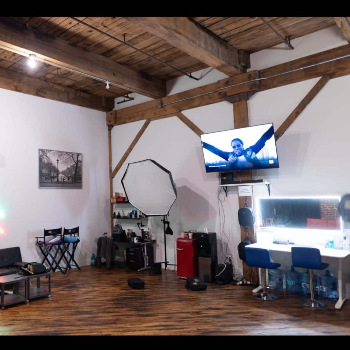 A stylistic photography studio with wooden beams, a couch, a makeup station, and a large monitor displaying an image on the wall.
