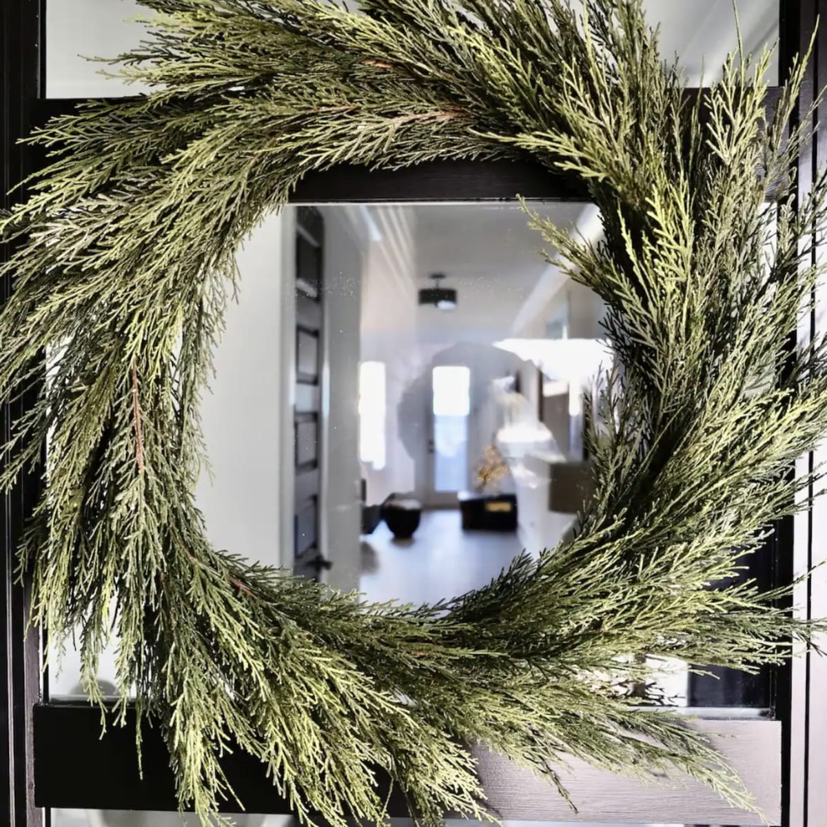A green wreath made of evergreen branches hangs on a glass door, reflecting the interior of a well-lit room.