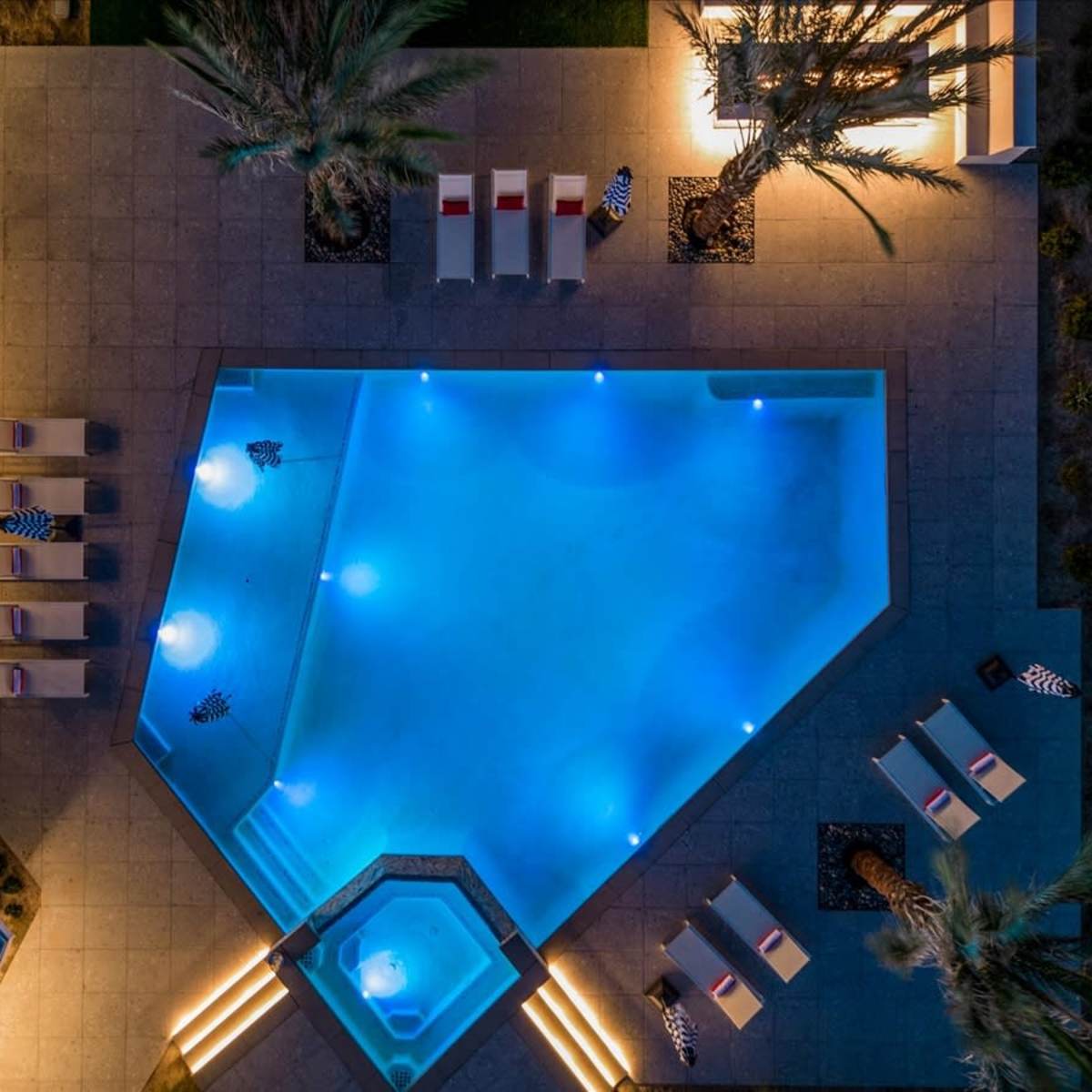 A geometric swimming pool illuminated by blue lights, surrounded by lounge chairs and palm trees.