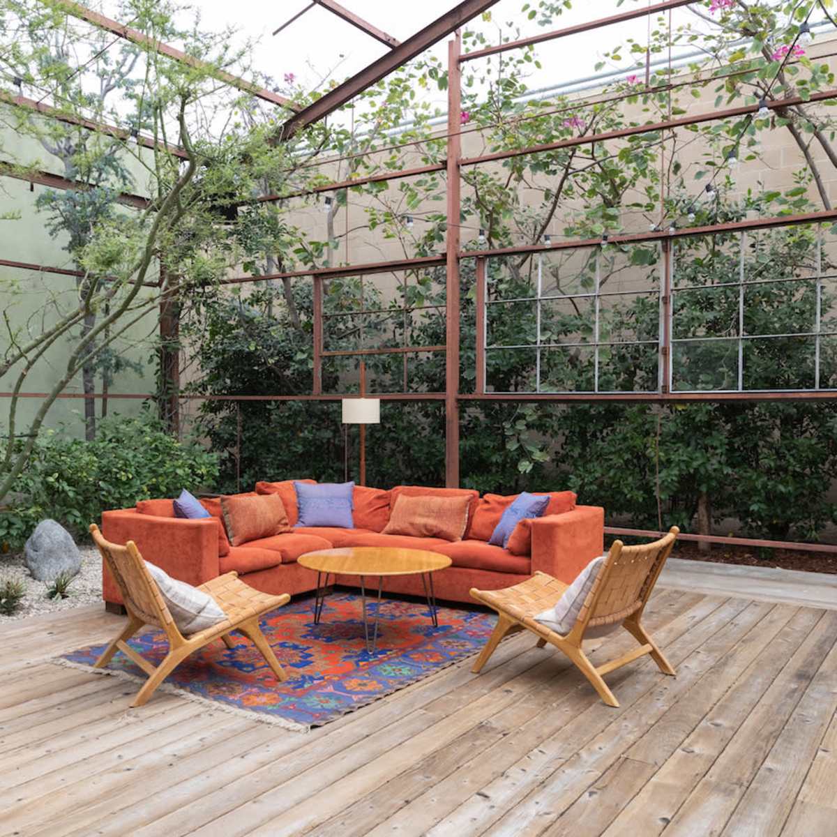A modern outdoor seating area features an orange sectional couch, a wooden coffee table, and two wicker chairs, surrounded by greenery and large windows.
