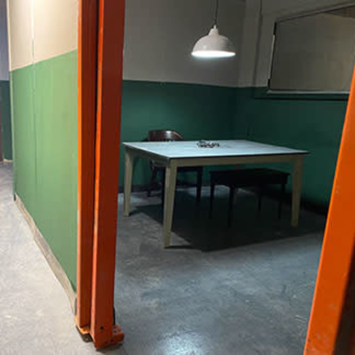 A sparse interrogation room with a table and two chairs, illuminated by a hanging light, bordered by green walls.