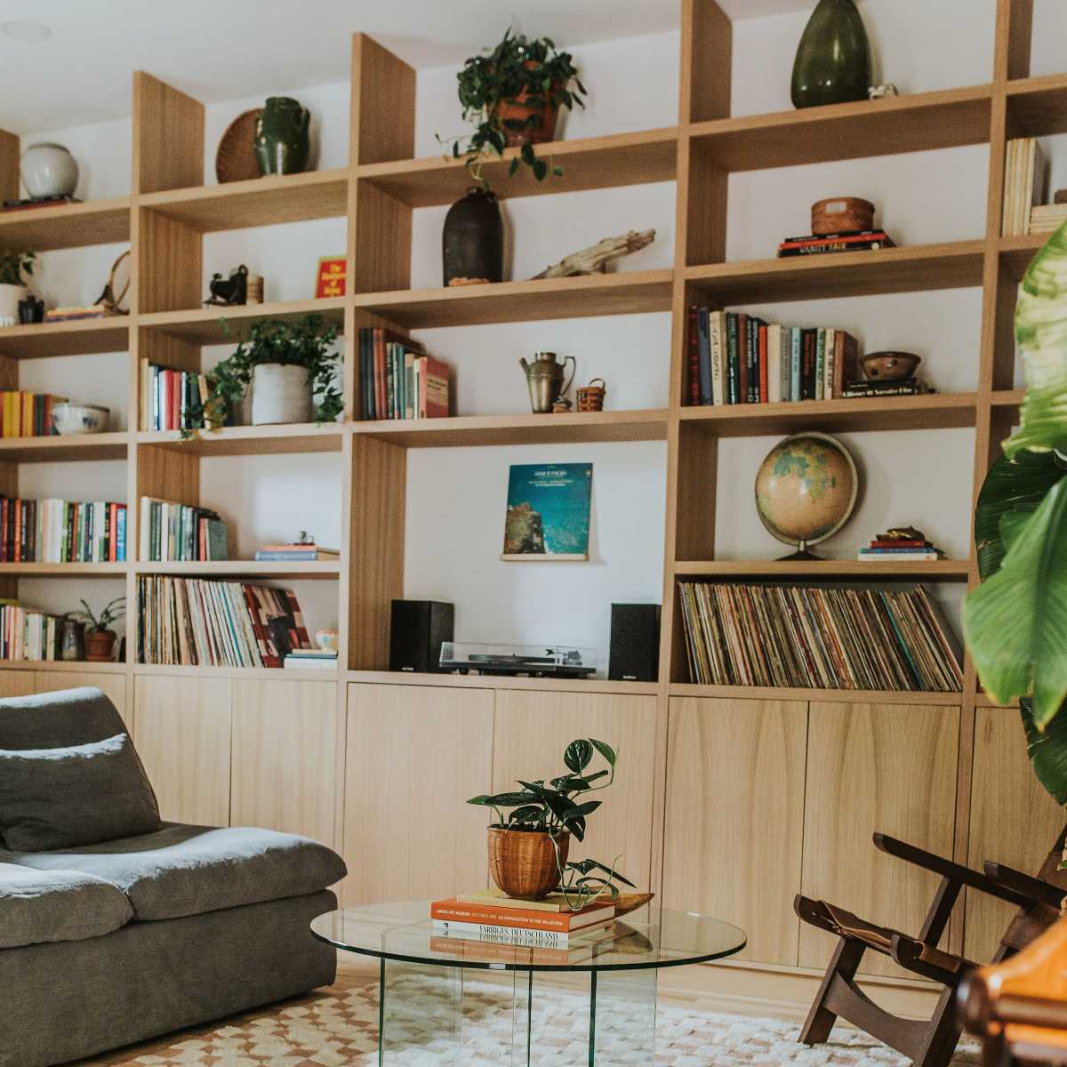 A cozy living room features a gray sofa, a glass coffee table with stacked books, and floor-to-ceiling shelves filled with plants and various decorative items.