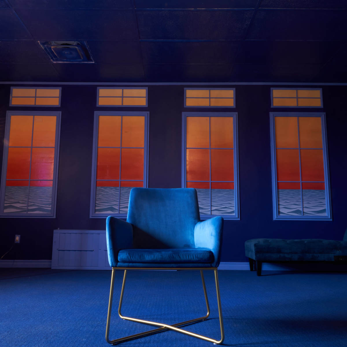 A blue chair sits in a dimly lit room with patterned walls and large windows displaying a colorful sunset.