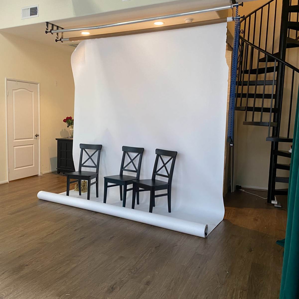 A photography studio featuring three black chairs in front of a white backdrop, with a spiral staircase and wooden floor visible in the background.