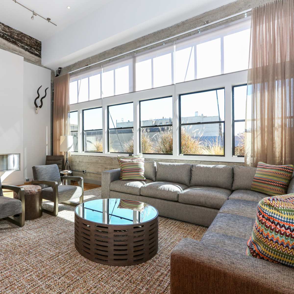 A modern living room with large windows, a gray sofa, a round coffee table, and decorative elements like animal horns on the walls.