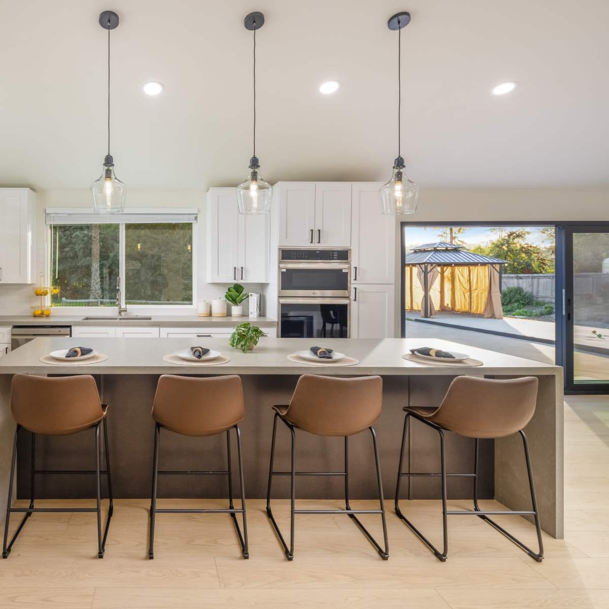 A modern kitchen features a concrete island with four brown bar stools, stainless steel appliances, and bright overhead lighting.