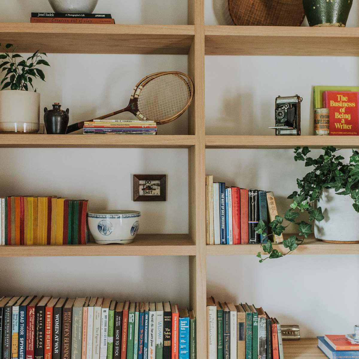 A wooden bookshelf displays a variety of books, plants, and decorative items, including a vintage camera and a tennis racket.