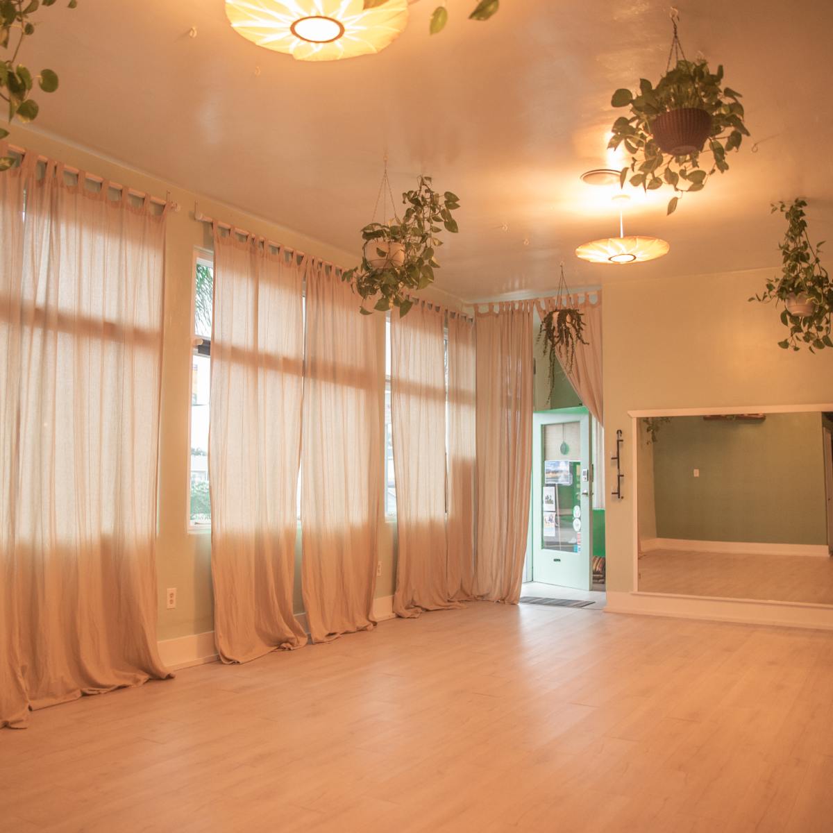 A spacious room with large windows, light-colored wooden flooring, and sheer curtains, along with several hanging plants and a full-length mirror on one wall.