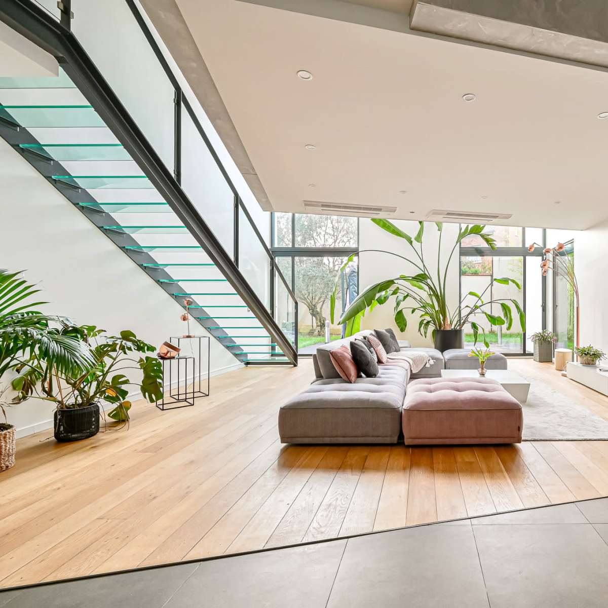 A modern living room features a large sectional sofa, a flat-screen TV mounted on a white wall, and tall windows with indoor plants surrounding a wooden floor.