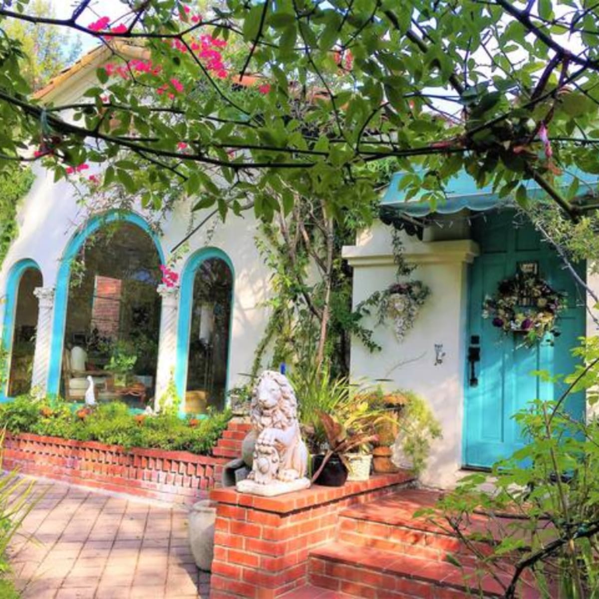 A house with an aqua door and arched windows is surrounded by greenery and features a stone lion statue on a brick pathway.