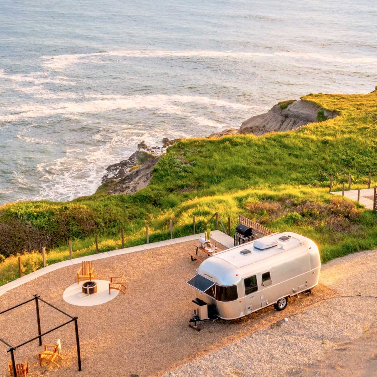 A silver Airstream trailer is parked on a gravel area overlooking the ocean, with a seating area and fire pit nearby.