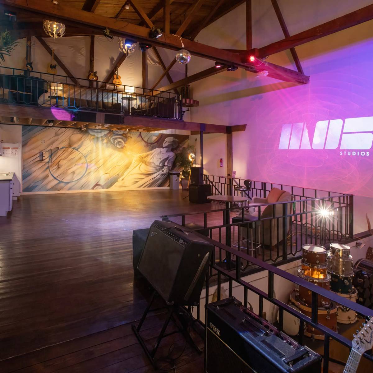 A spacious studio with wooden beams, a mural on the wall, musical instruments set up, and dim lighting featuring colored spotlights.