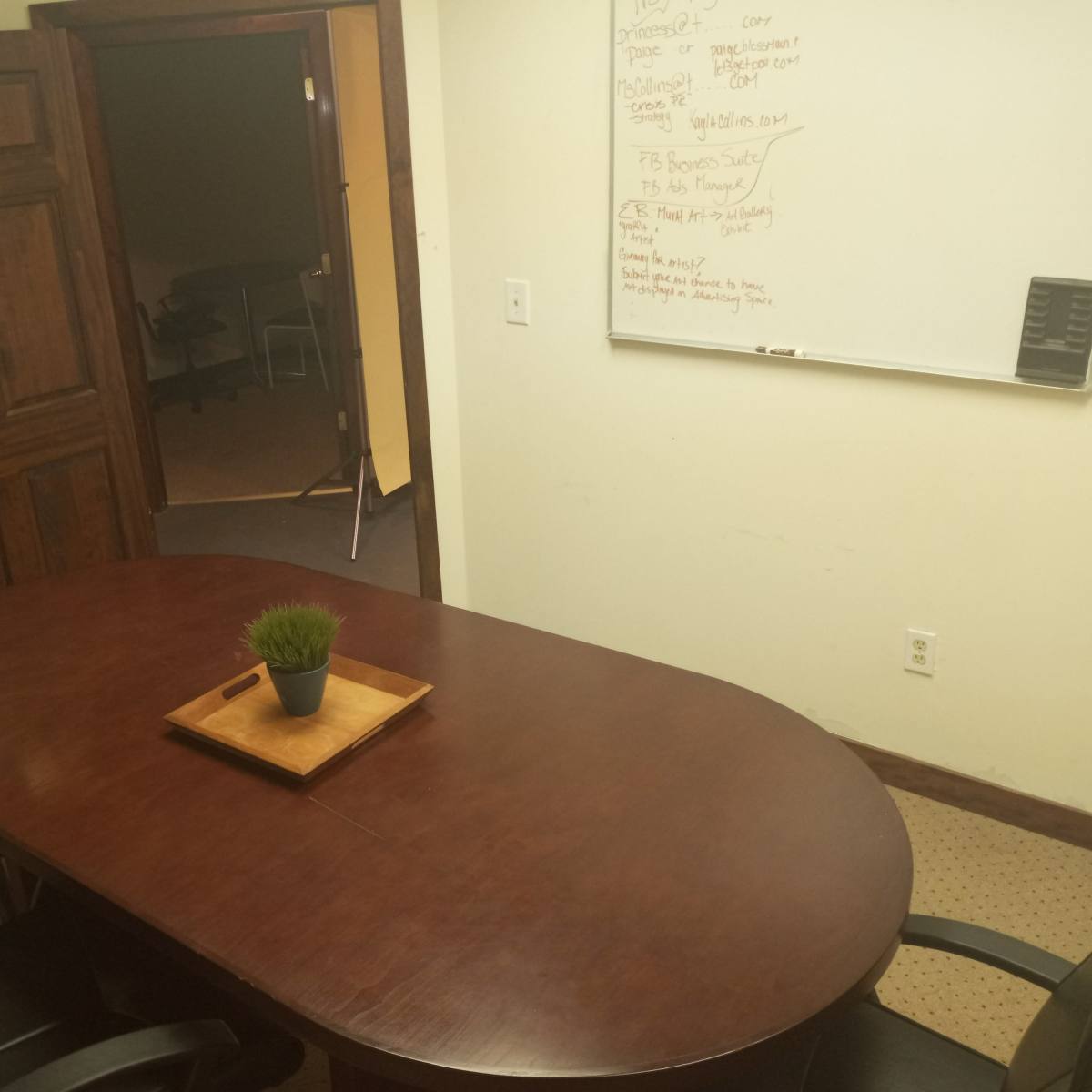 A conference room features a large wooden table surrounded by black leather chairs, with a small potted plant on the table and a whiteboard on the wall.