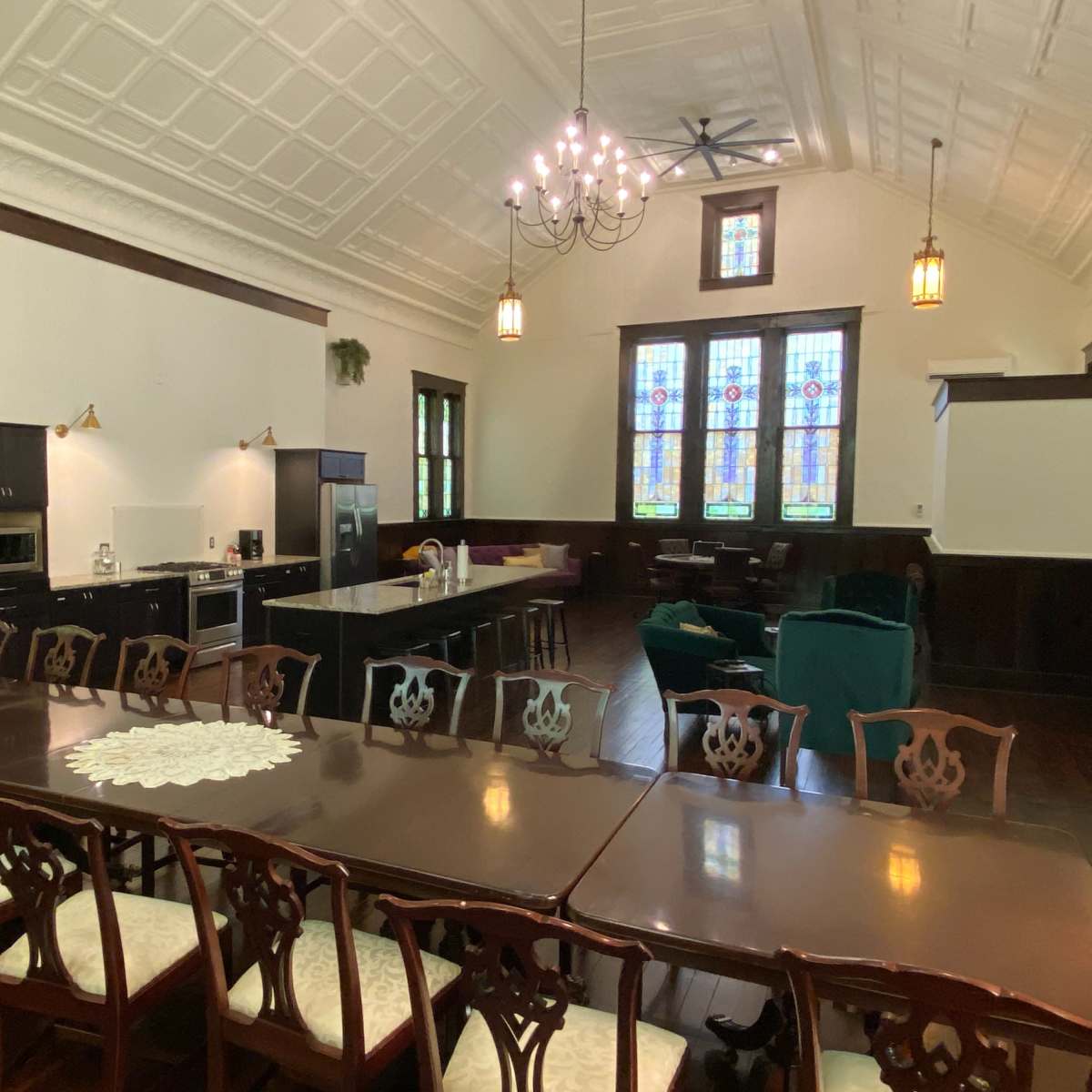 A spacious room with a long dining table, a modern kitchen area, and large stained glass windows.