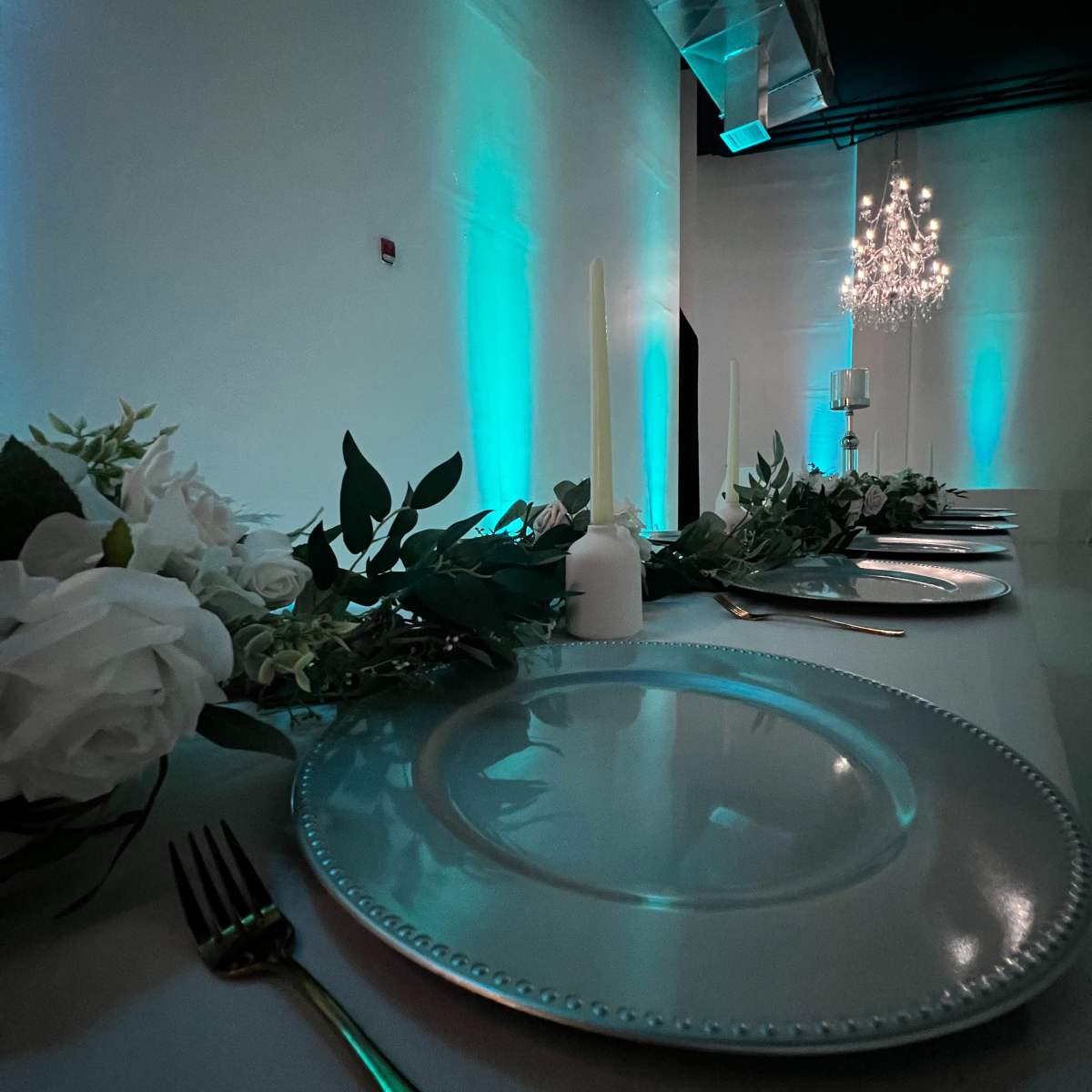 A long, elegantly set dining table is decorated with white plates, green foliage, and soft candlelight under teal lighting and a chandelier.