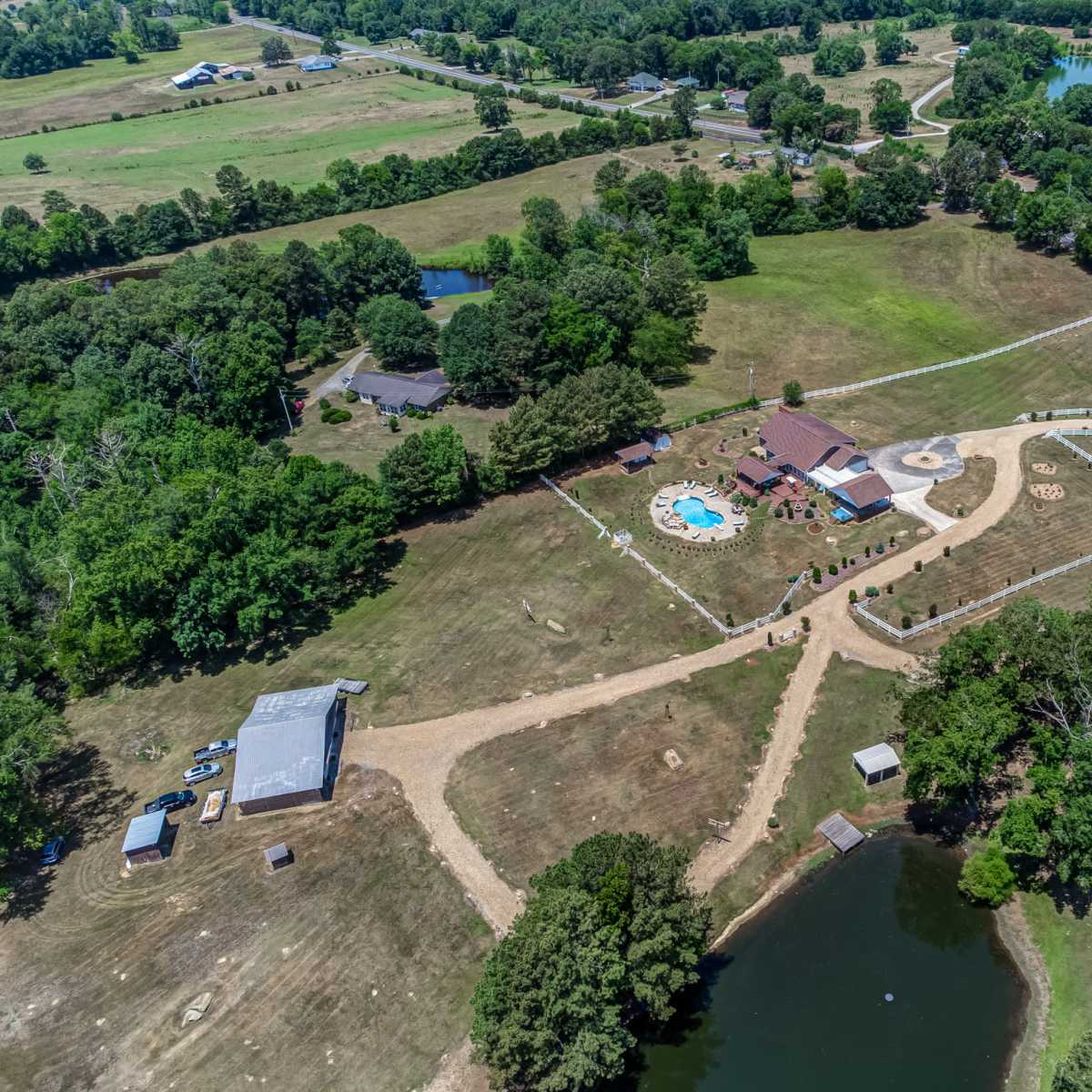 An aerial view of a rural property featuring a house with a pool, surrounded by landscaped grounds, a pond, and multiple outbuildings.