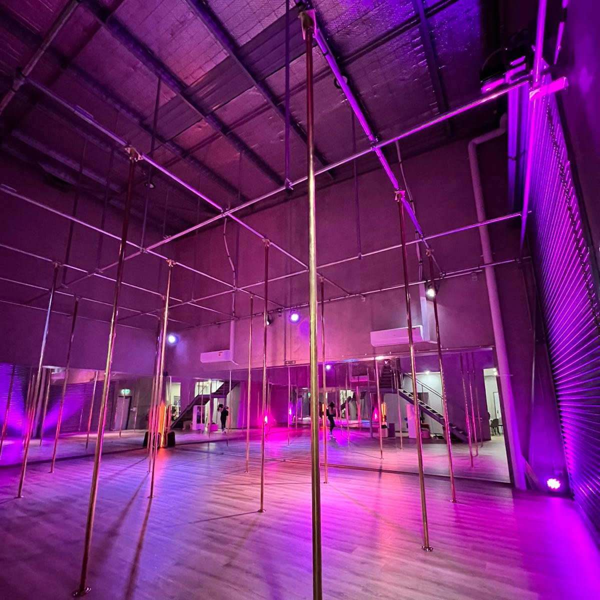 A spacious dance studio featuring several polished poles, illuminated by purple and pink lighting.