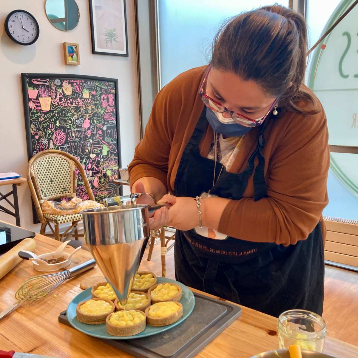 A woman wearing a mask is carefully piping filling onto small tarts in a kitchen setting.