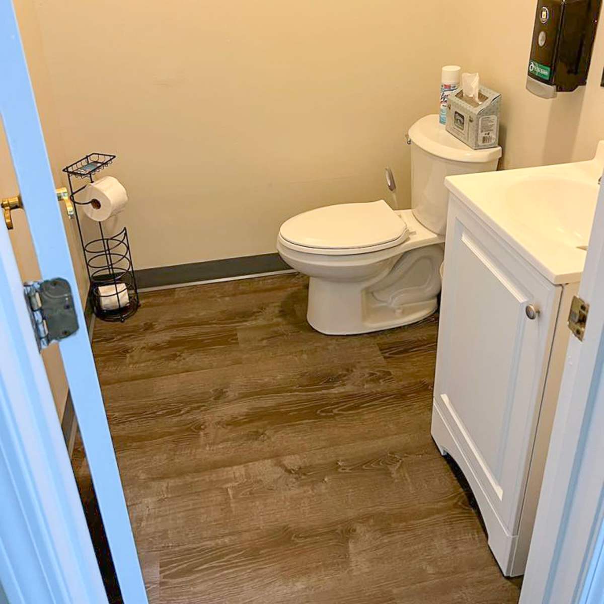 A small bathroom containing a toilet, a sink with a cabinet, a toilet paper holder, and a hand sanitizer dispenser on the wall.