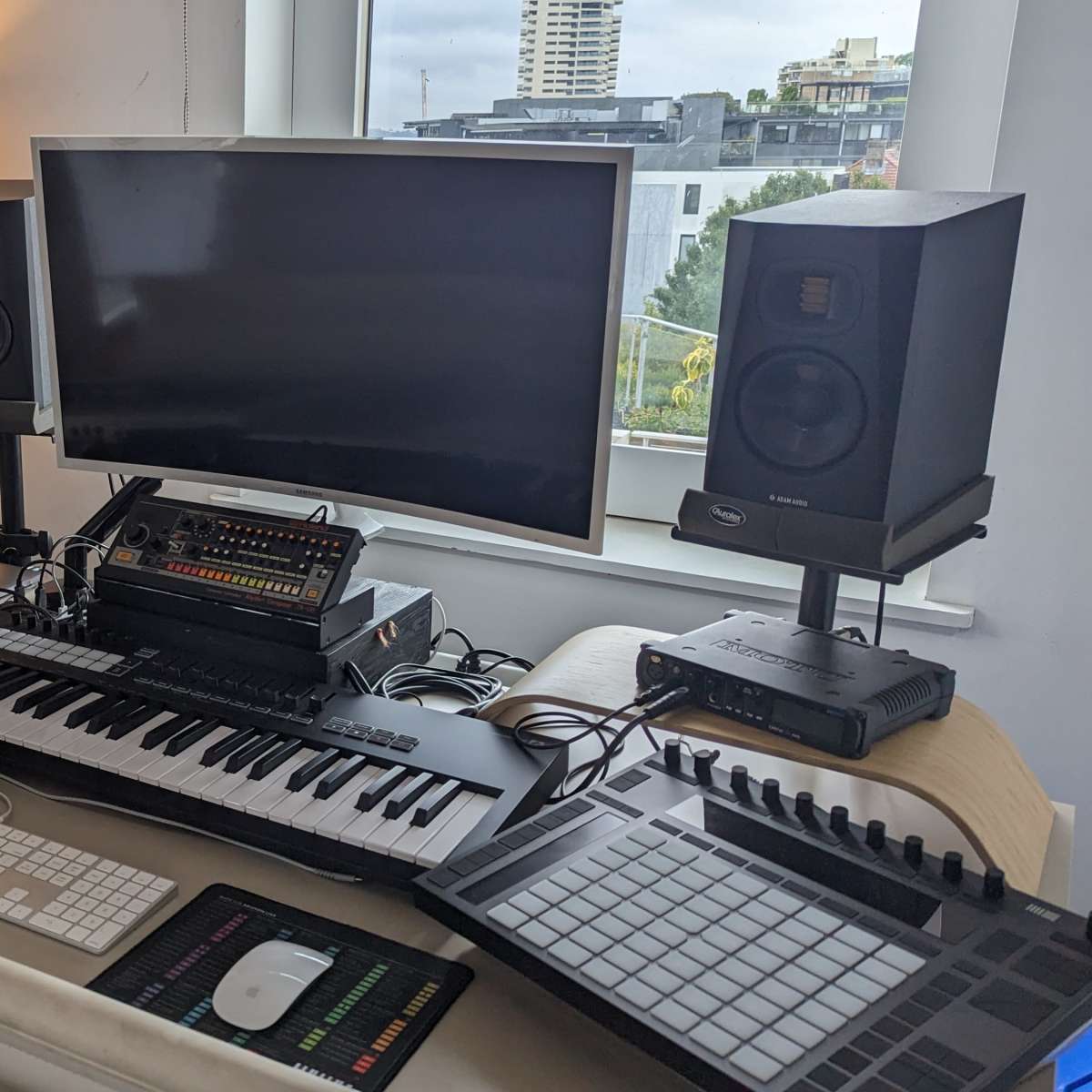 A modern music production workspace featuring a computer monitor, audio speakers, a keyboard, and various electronic instruments, with guitars positioned in the background.
