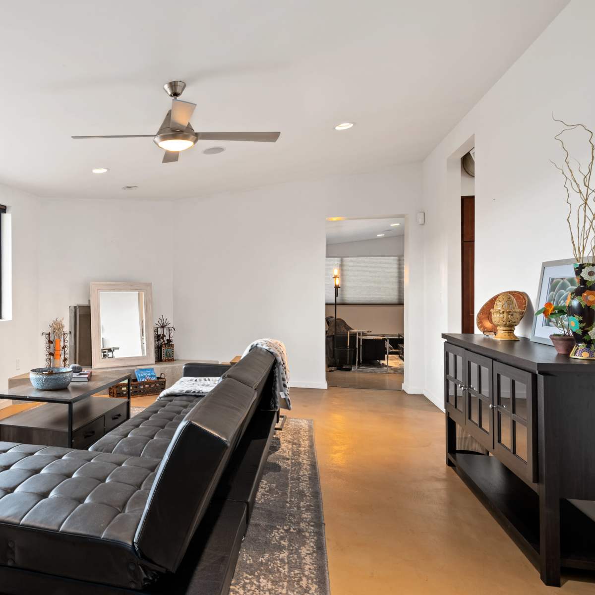 A modern living room featuring a black leather sofa, a coffee table, and large windows with a view of greenery outside.