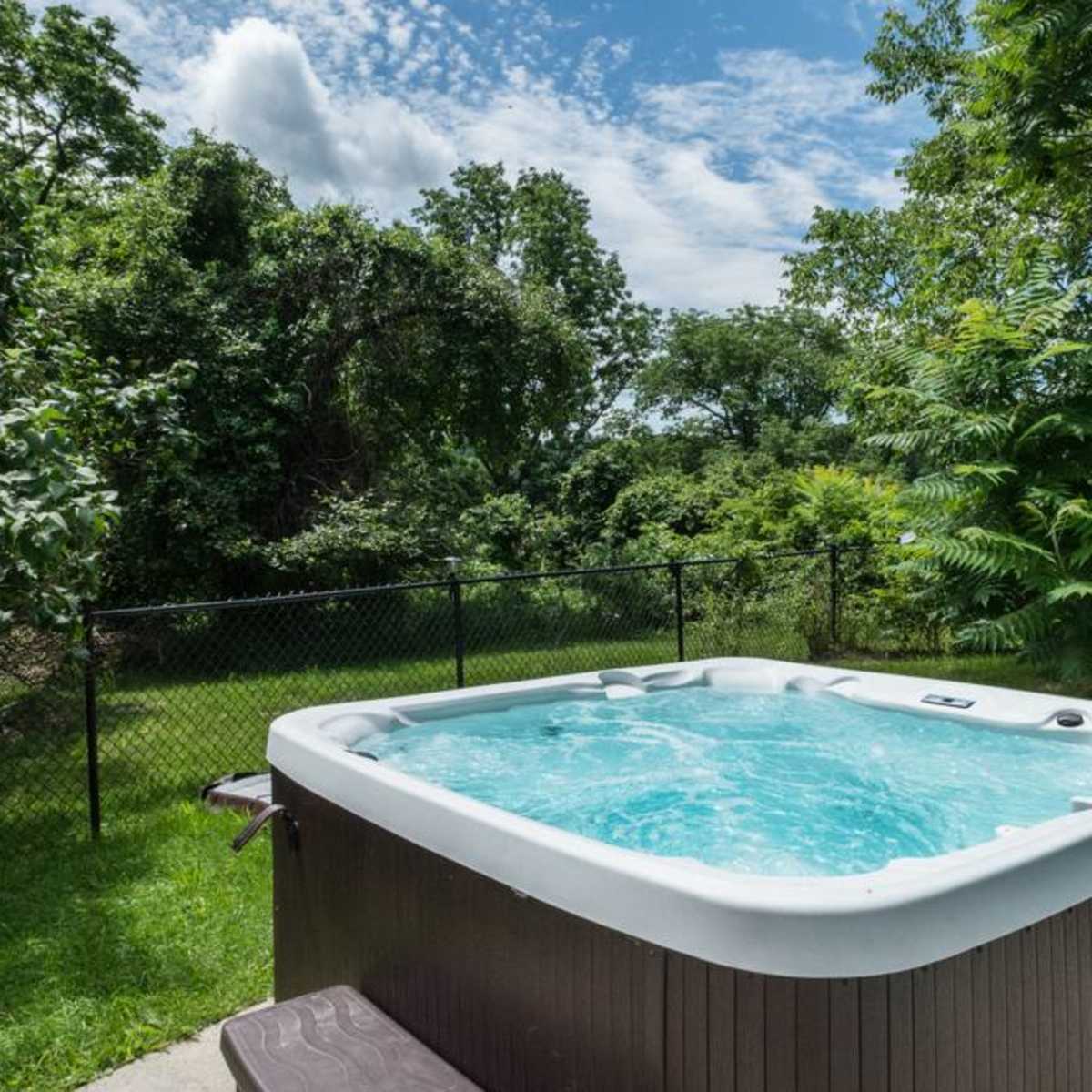 A hot tub sits on a patio surrounded by green trees and bushes, with a fenced yard in the background.