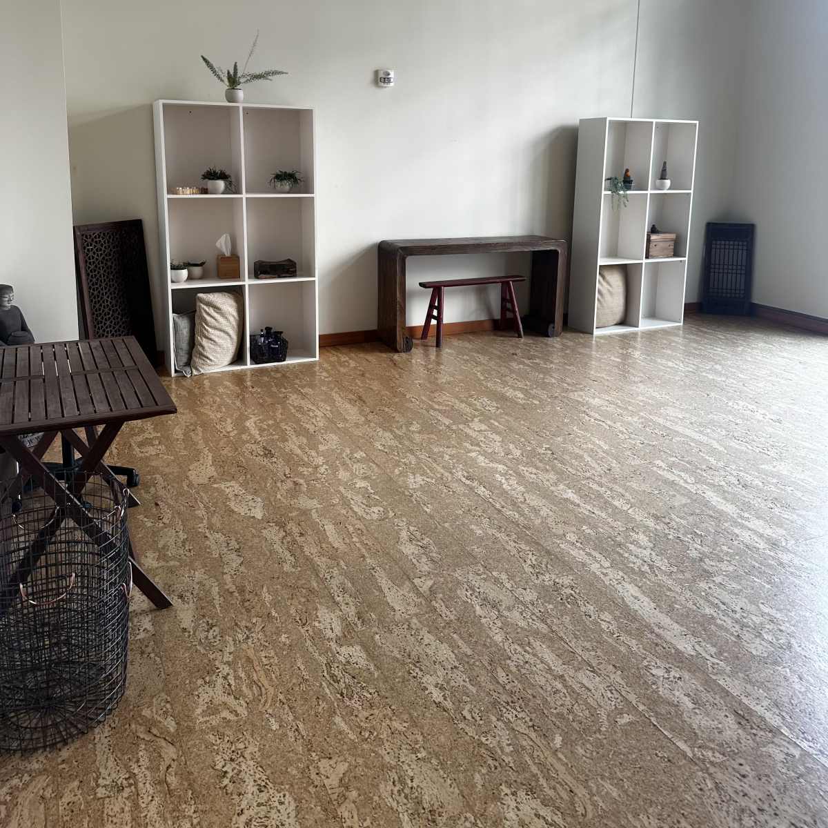 A spacious, minimally furnished room with two white shelving units, a wooden bench, a small table, and a wicker basket on the floor.